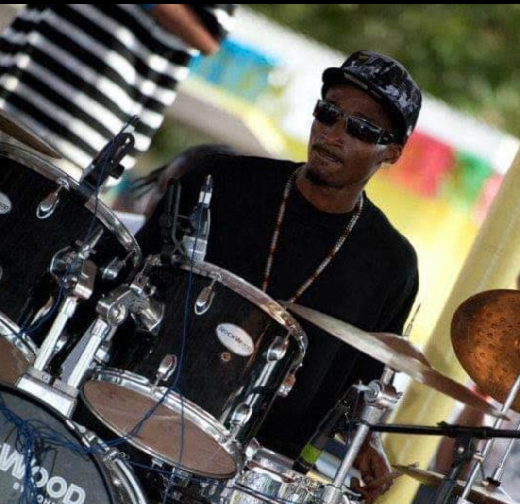 Man playing drums outdoors with sunglasses and a cap.