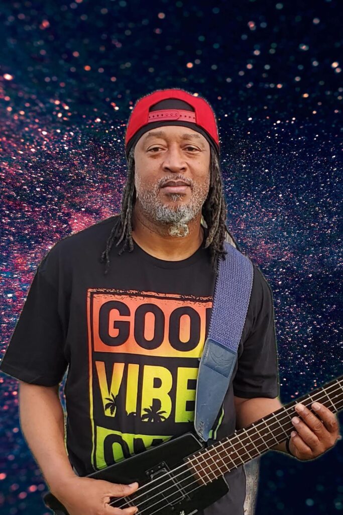 Man with dreadlocks wearing a red cap and a "Good Vibes" shirt, holding a guitar.